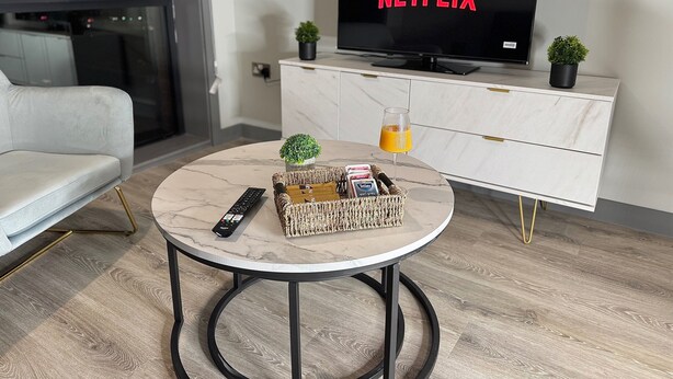 Ein modernes Wohnzimmer mit einem runden Marmortisch, einer Couch, einem Fernseher, der das Netflix-Logo zeigt, einem Glas Orangensaft und einem Korb mit Büchern und einer Fernbedienung.