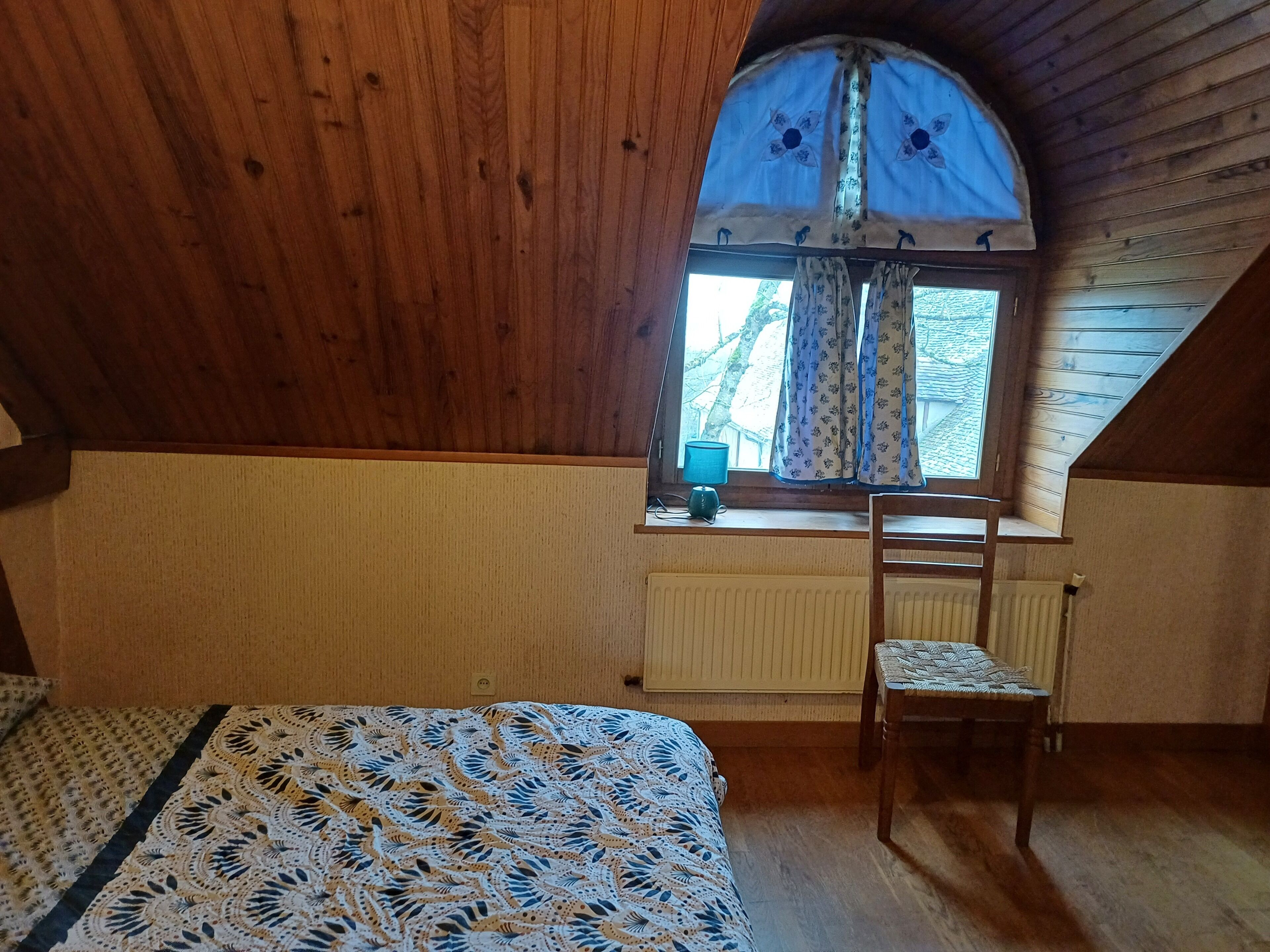 Een knus ingerichte kamer met houten plafond, een raam met bloemen gordijnen, een radiator, een bed met een beddegoed met dessin, en een houten stoel.