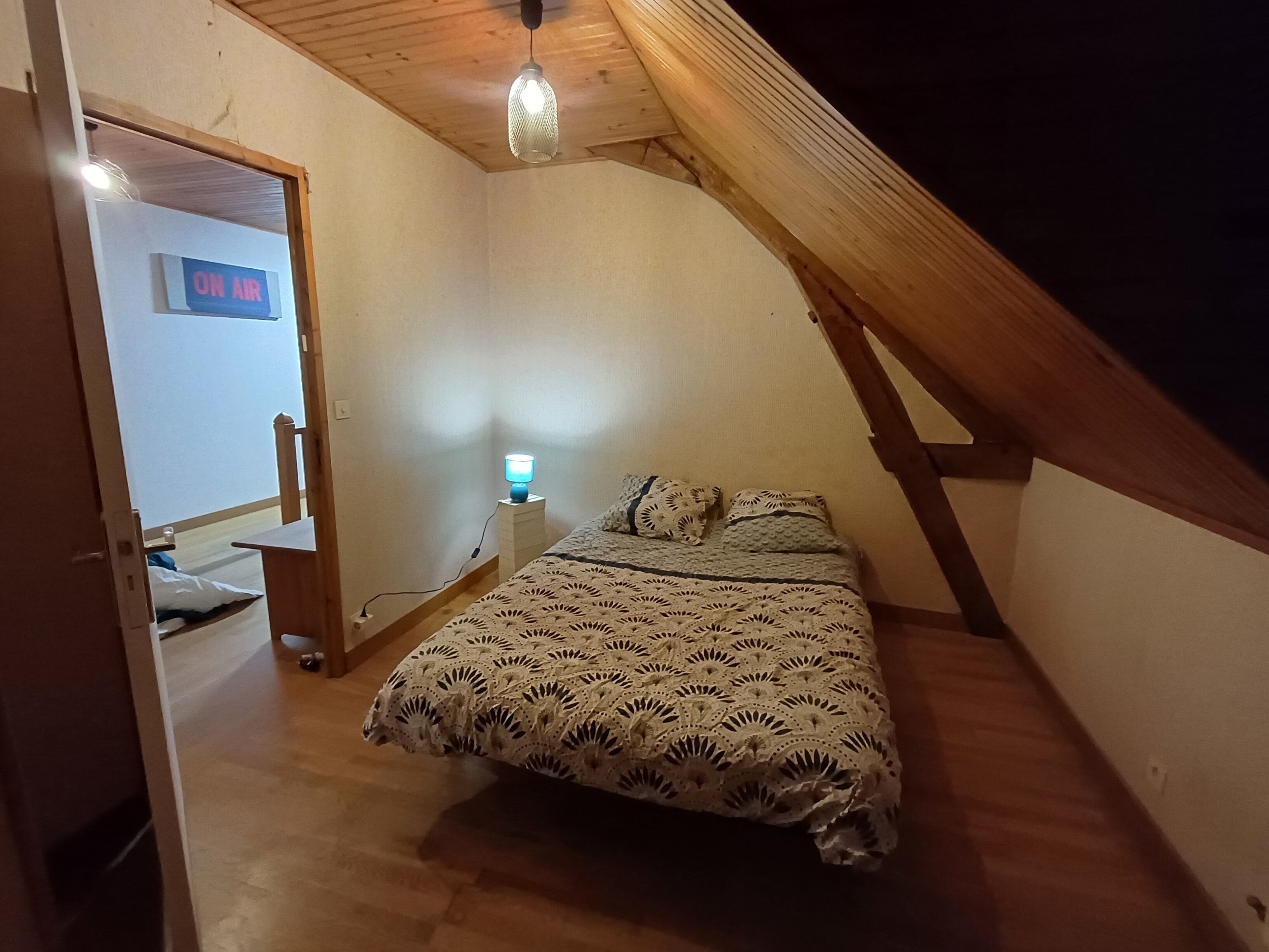 A small, cozy attic bedroom with a sloped ceiling, a bed with a patterned blanket, a bedside lamp, and a sign indicating the room is on air.
