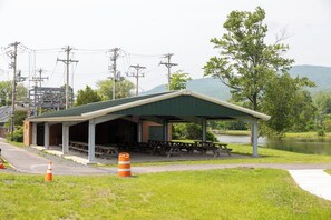 A covered picnic area with benches and picnic tables, surrounded by a grassy area and a body of water.