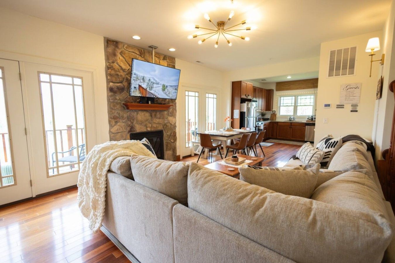 A spacious living room with a large sectional sofa, a fireplace, a television mounted on a stone wall, and a dining area with a table and chairs.