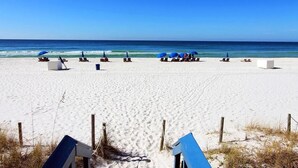 Ein Strand mit weißem Sand und klarem, blauem Wasser. Mehrere blaue Sonnenschirme und Liegestühle sind vorhanden.