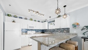 A modern kitchen with white cabinets, a granite island with bar stools, and decorative plates on the wall. - Gulf Place Caribbean 313- Sunsets on 30A (Santa Rosa Beach)