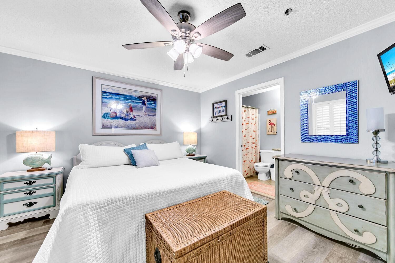 A bedroom with a bed, nightstands, a dresser, a ceiling fan, a painting, a mirror, and a wicker basket.