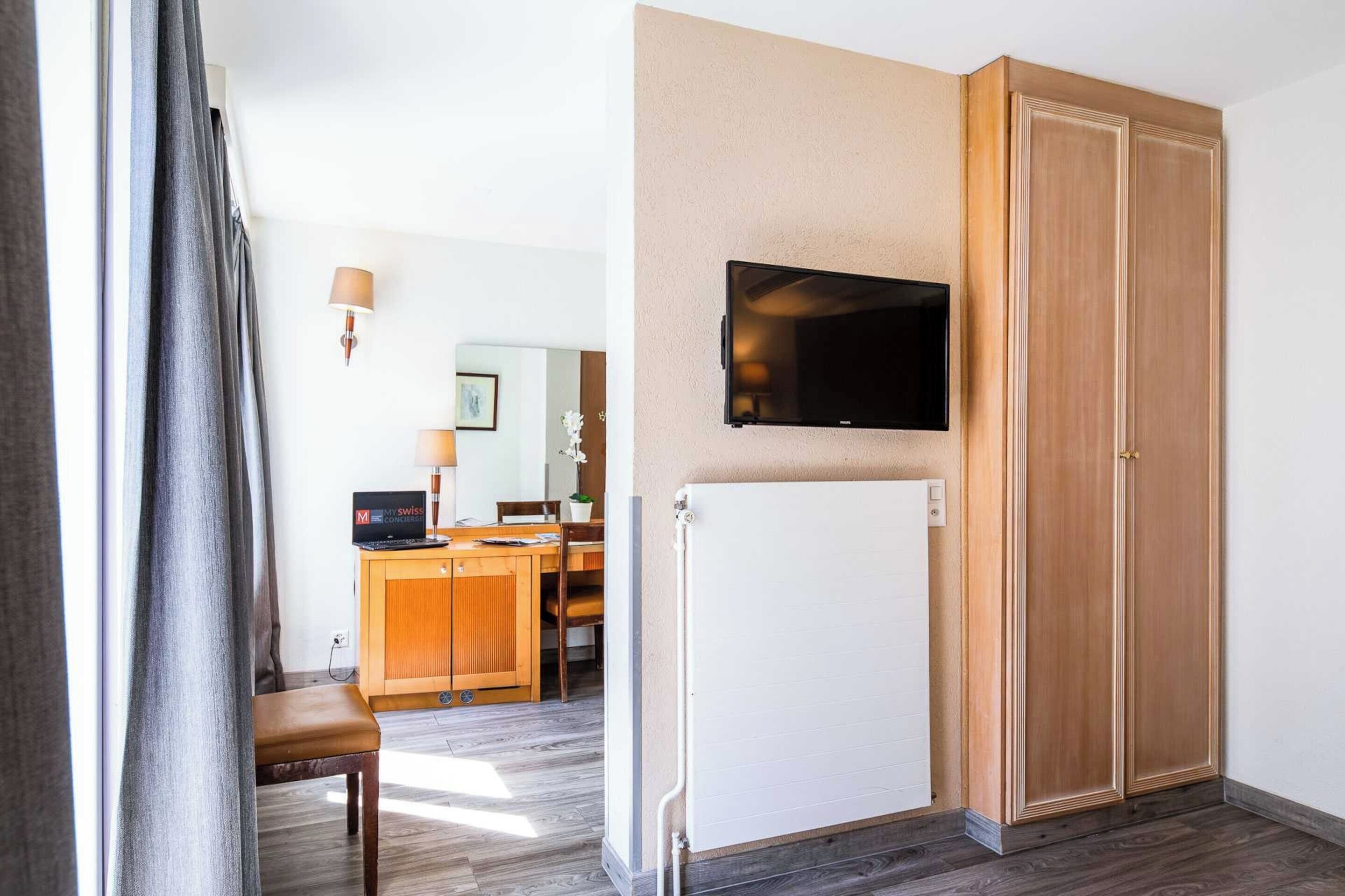 Ett hotellrum med en garderob i trä, en väggmonterad TV, ett skrivbord med en dator och ett vitt element.
