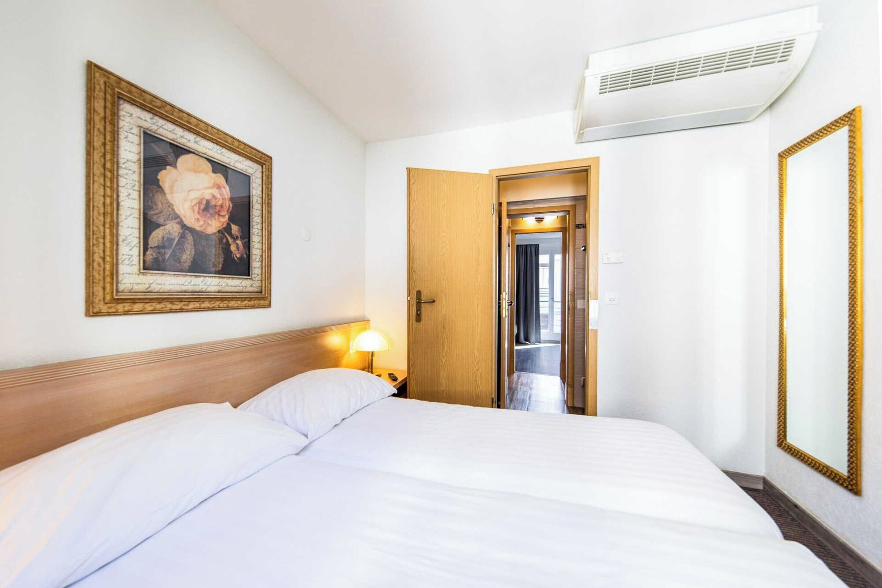 Une chambre d’hôtel avec un grand lit, un tableau encadré au mur, une porte donnant sur une autre pièce et un climatiseur fixé au mur.