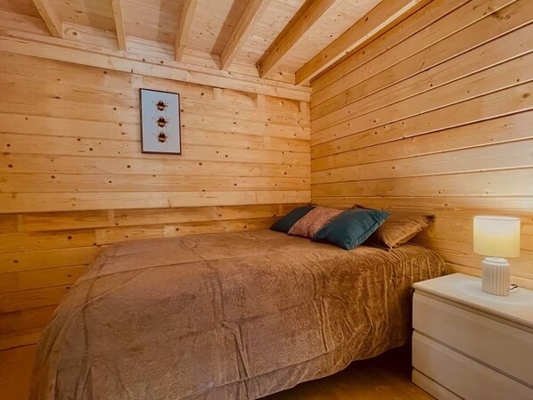 Ein Zimmer in einer Holzhütte mit einem Bett, Kissen, einem Nachttisch und einem Bilderrahmen mit Bienen an der Wand.