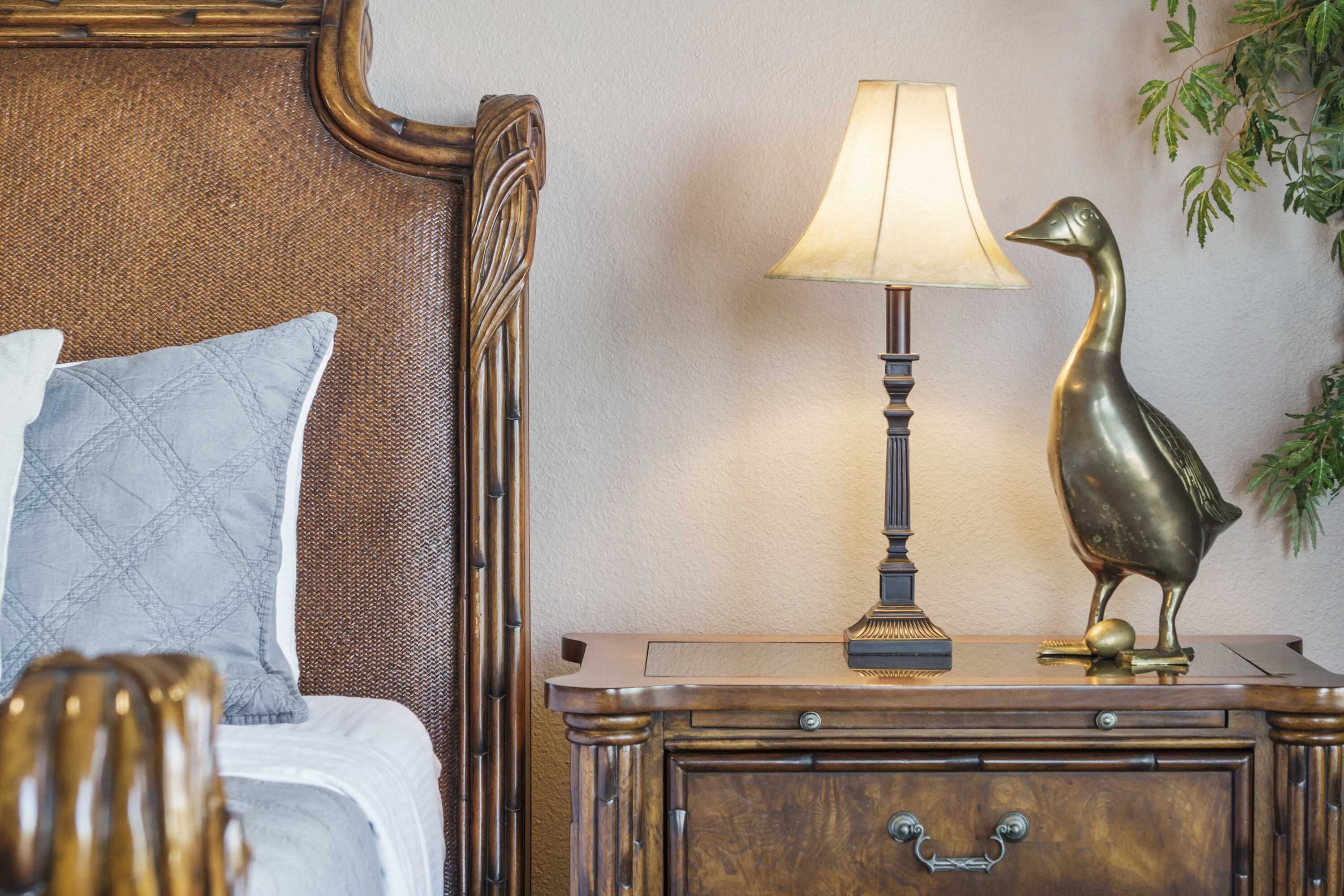 A neatly made bed with a headboard, a bedside lamp, a decorative bird statue, and a nightstand with drawers.