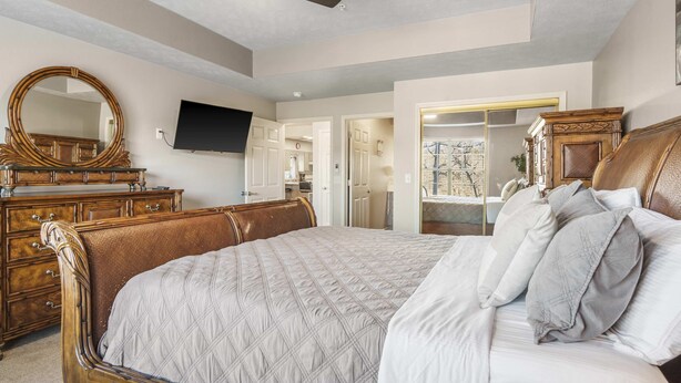 Une chambre avec un grand lit, une tête de lit en bois, un ventilateur de plafond, un téléviseur fixé au mur et une commode avec un miroir.