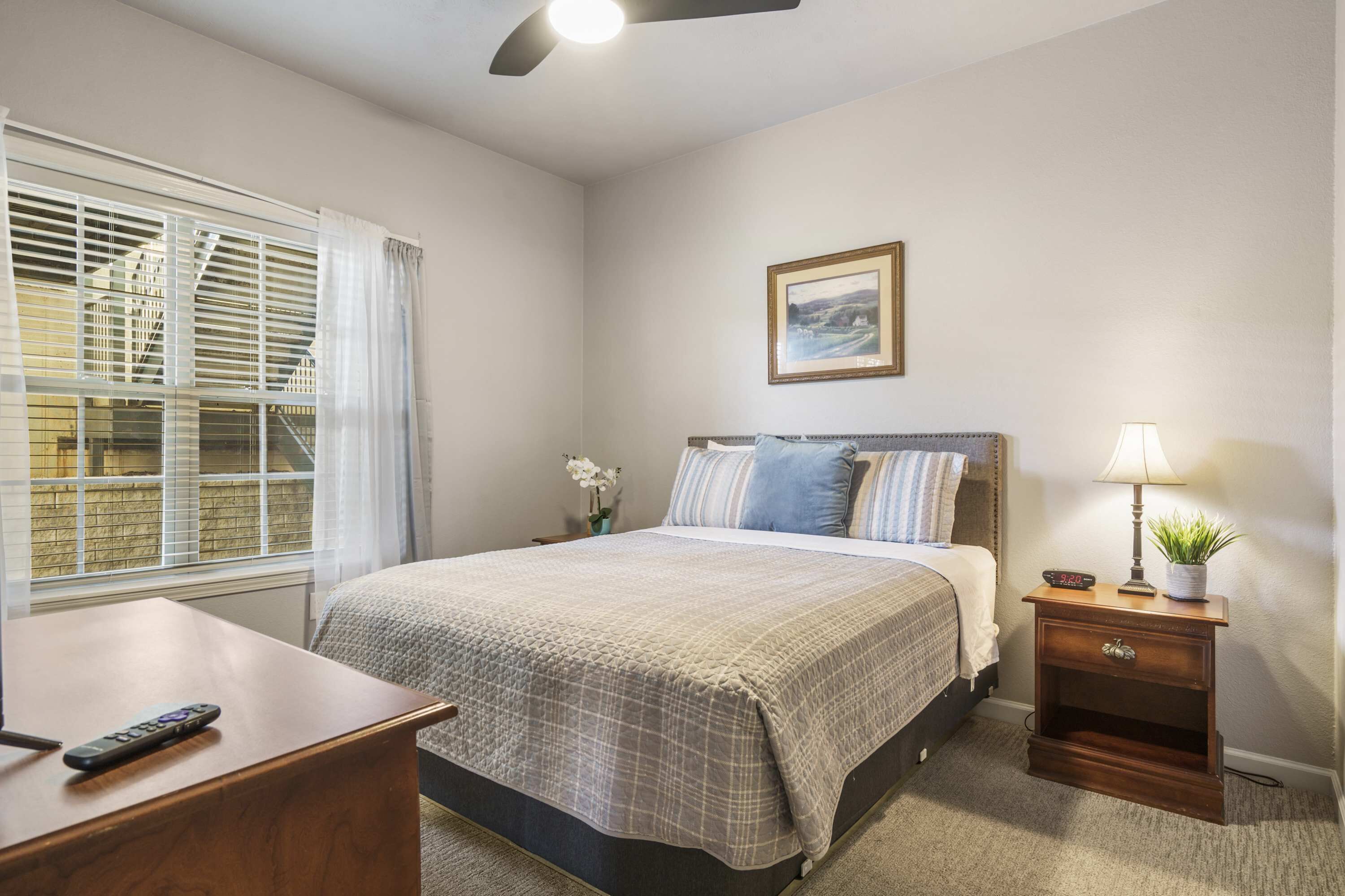 A bedroom with a bed, bedside table, nightstand, and a window with blinds.