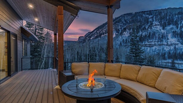 A wooden deck with a round table featuring a fire, a cushioned sofa, and a view of snow-covered mountains. - Alpine Luxury | Engineer Mtn View | Pool & Hot Tub (Durango)