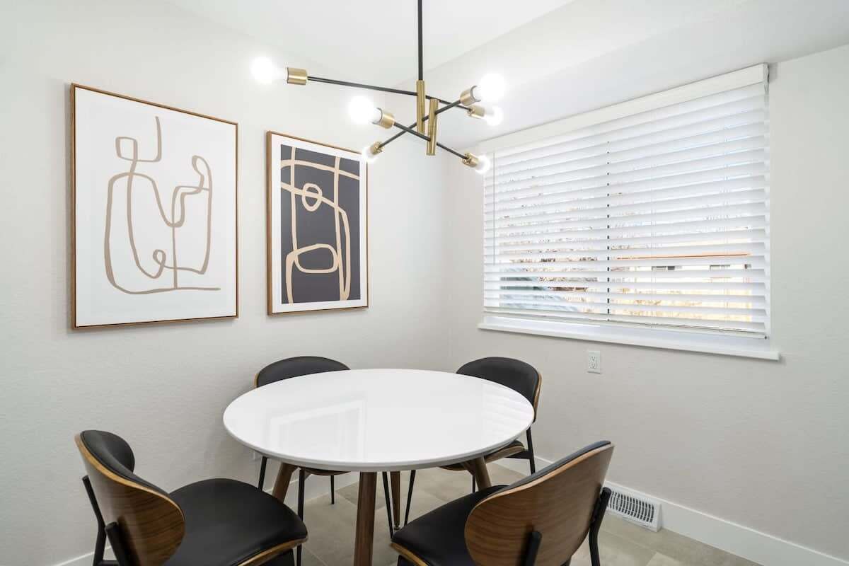 Een eethoek met een ronde tafel en zes stoelen, twee ingelijste abstracte kunstwerken aan de muur, en een raam met jaloezieën.