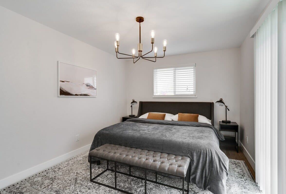 Quarto com cama grande, um banquinho, um candelabro, janela com persianas e um quadro na parede.