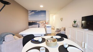 Une chambre d’hôtel avec un lit, une table à manger dressée avec des assiettes et des tasses, une télévision et une vue sur la plage, visible à travers une grande fenêtre.