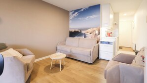 Un salon avec un canapé beige, une petite table ronde et un coin cuisine doté de meubles blancs et d’un plan de travail.