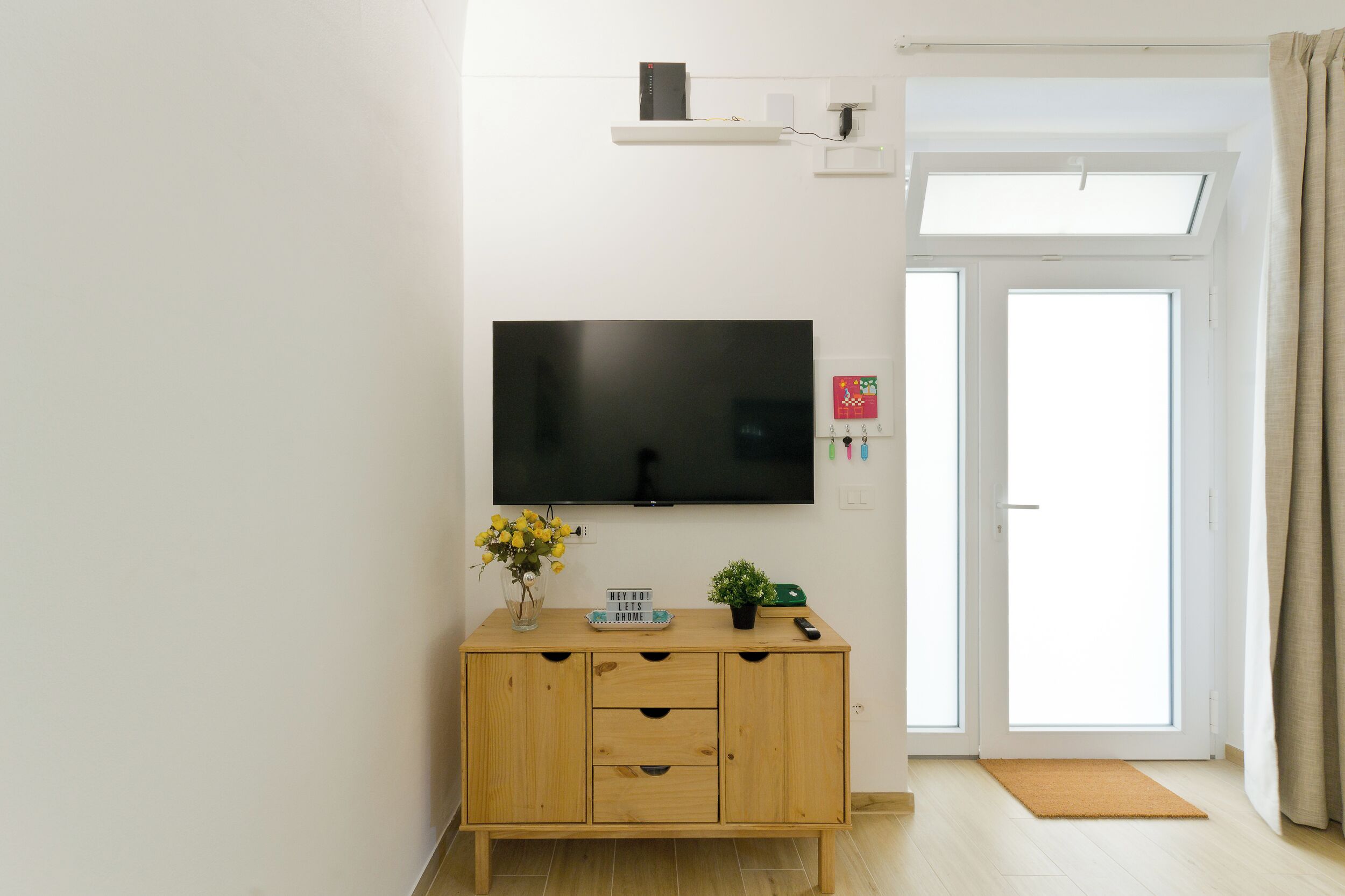 Un salon moderne avec un téléviseur à écran plat fixé au mur, un meuble en bois orné d’un vase de fleurs et une porte-fenêtre coulissante donnant sur un espace extérieur.