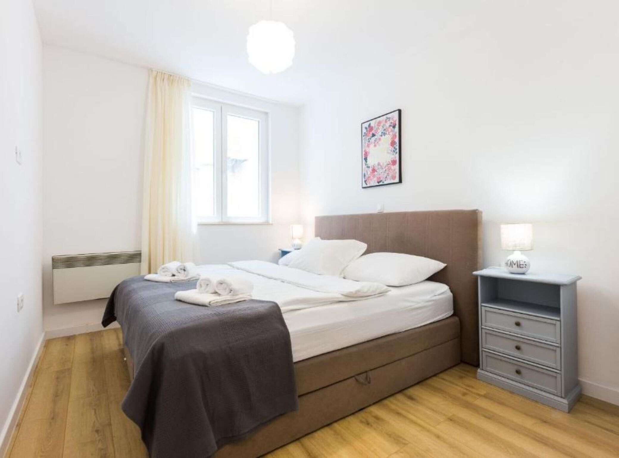Une chambre à coucher comprenant un lit, des tables de chevet, un radiateur, une fenêtre avec des rideaux et un tableau accroché au mur.