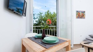 Un coin repas avec une table en bois, des assiettes vertes et un vase avec une fleur. Un téléviseur est fixé au mur, et on aperçoit un balcon avec une rambarde et de la verdure.
