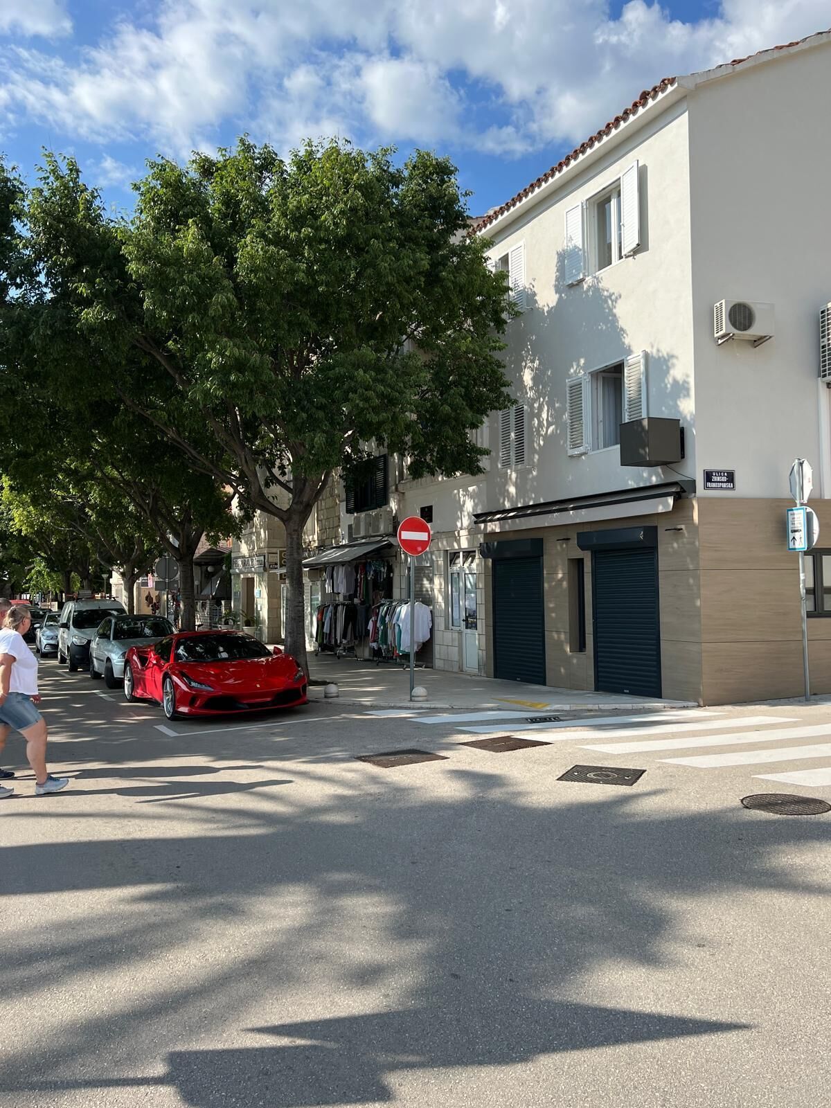 Una strada con un'auto sportiva rossa parcheggiata a lato, un attraversamento pedonale e edifici con negozi.