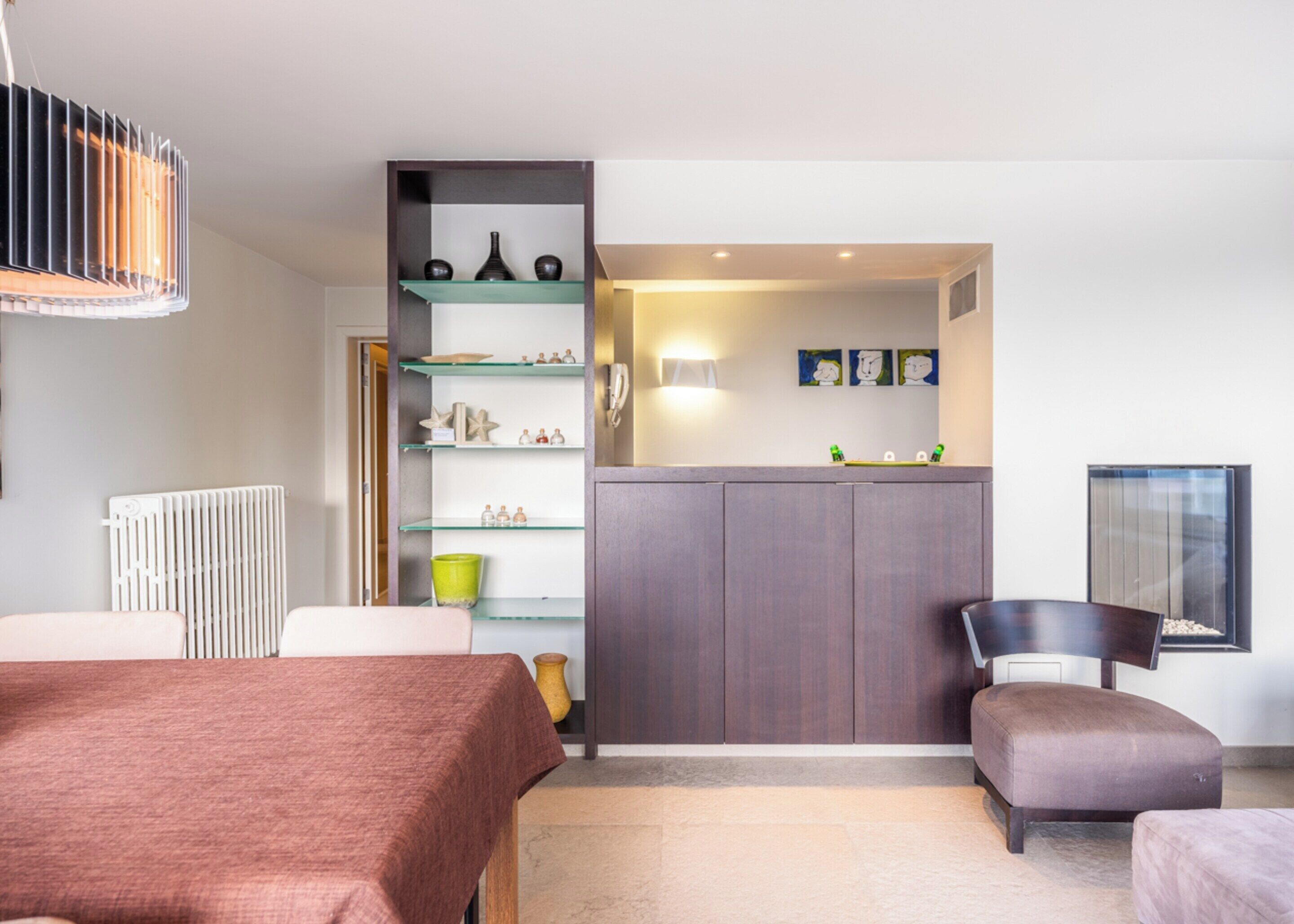 A modern bedroom with a bed, a small seating area, a built-in shelf unit, and a radiator.