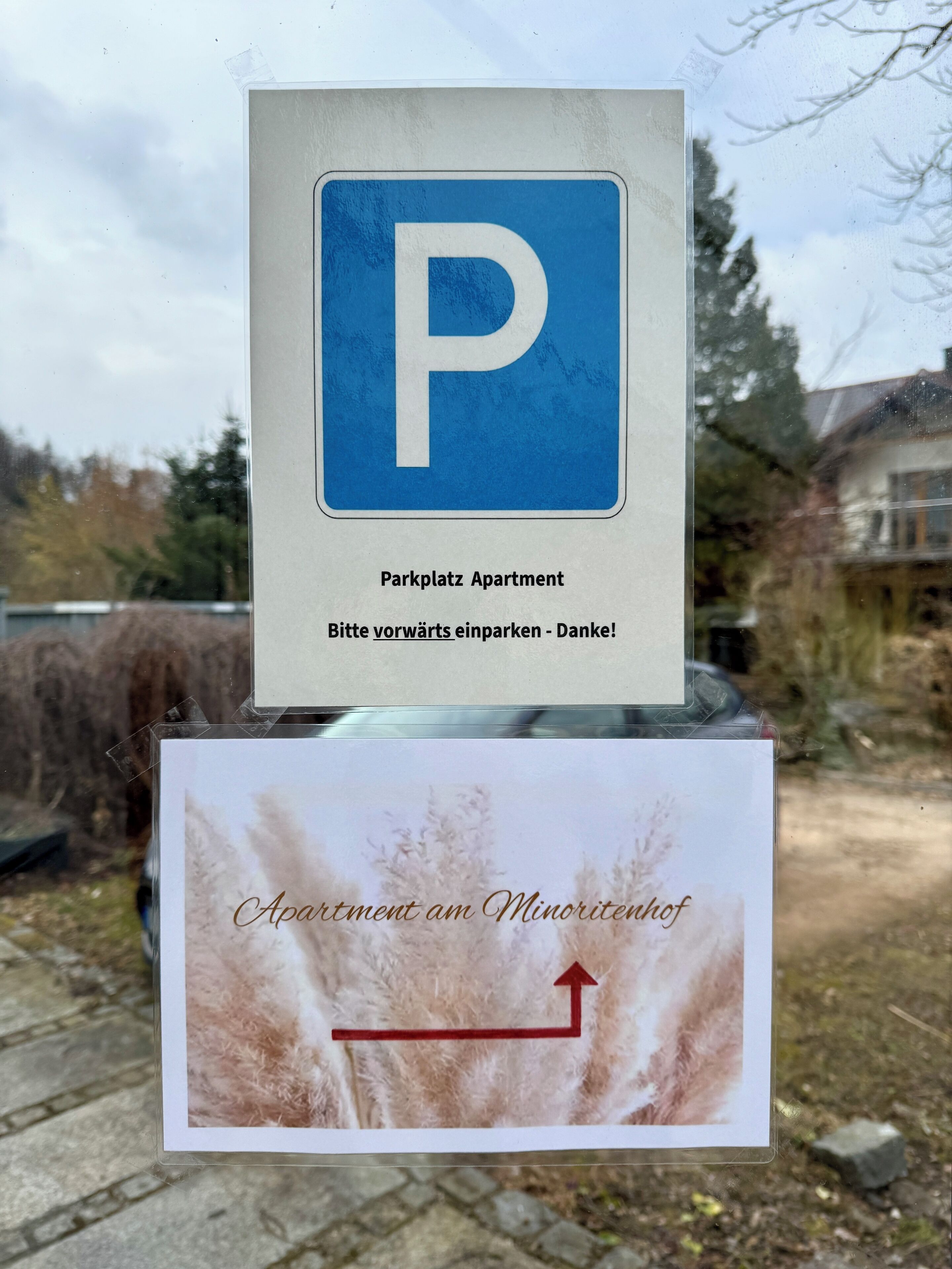 A parking sign with a blue 'P' and text indicating 'Parkplatz Apartment' and 'Bitte vorswarts einparken - Danke!' with a directional arrow pointing to the left.