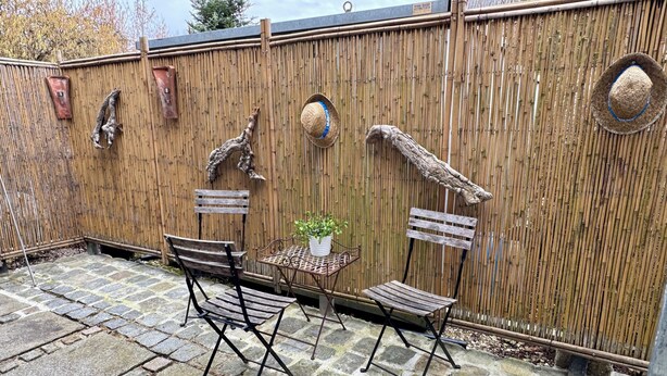 Un patio con pavimento in pietra, due sedie di legno, un tavolino con una pianta e un recinto di bambù con elementi decorativi.