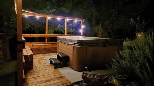 Ein Whirlpool auf einer Holzterrasse mit einer Pergola darüber, umgeben von Topfpflanzen und einer Holzbank.