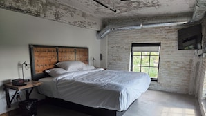 A bedroom with a large bed, a wooden headboard, a nightstand with a lamp, and a window with a view of greenery. - The Mill Hotel (Fergus Falls)