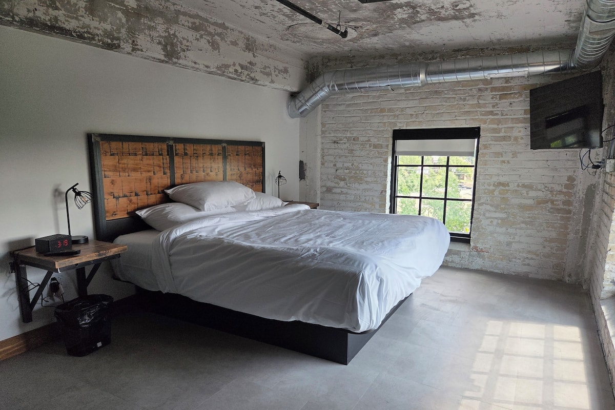 A bedroom with a large bed, a wooden headboard, a nightstand with a lamp, and a window with a view of greenery.