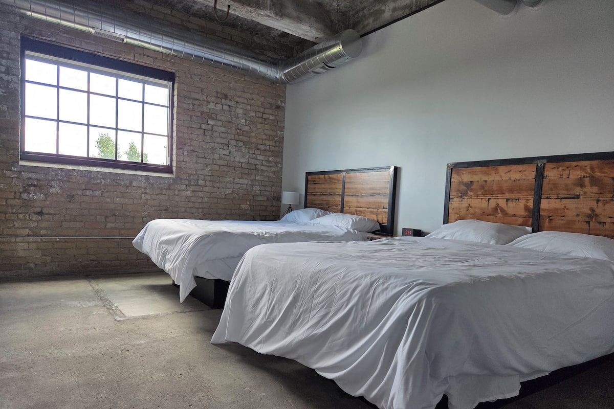 A room with two beds, a brick wall, a large window, and a concrete floor.