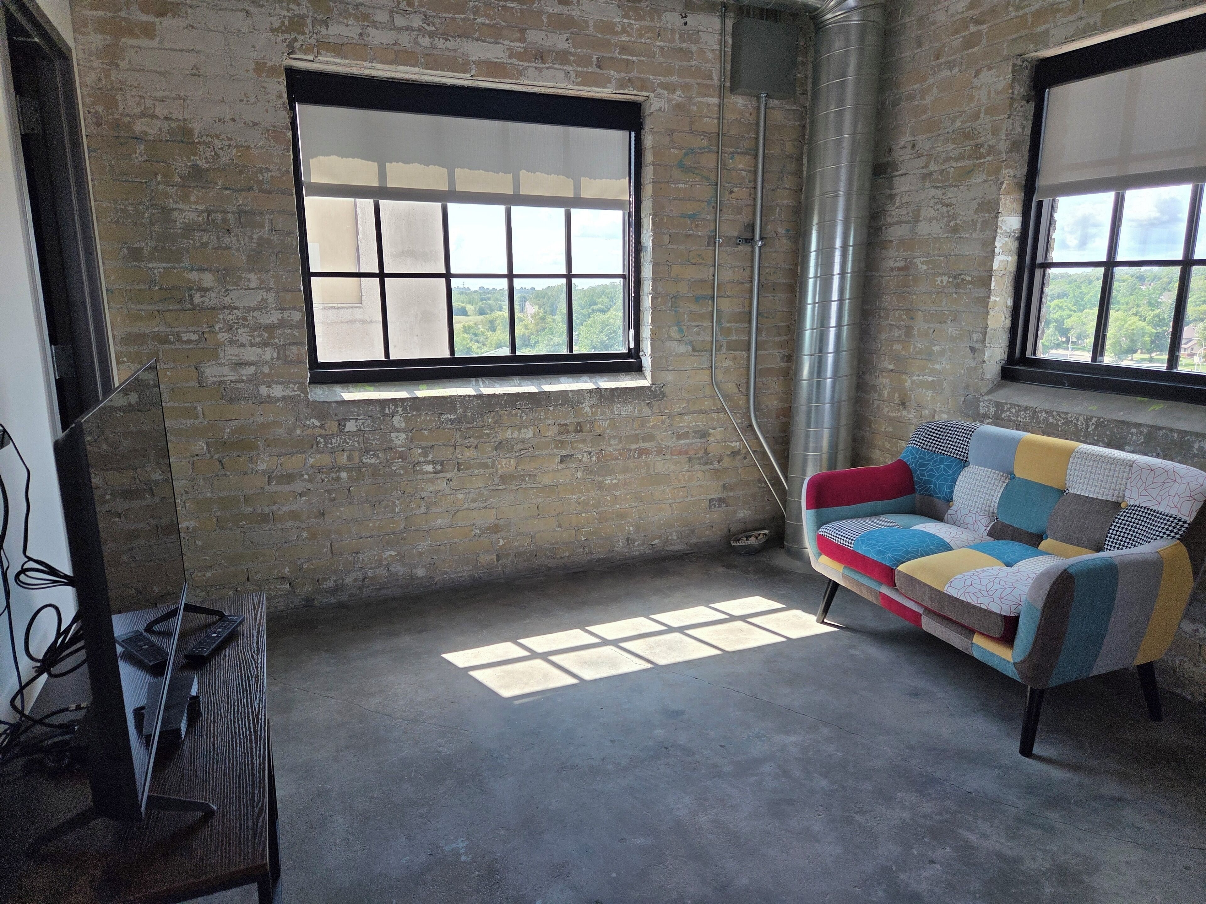 A room with exposed brick walls, a large window with a view of greenery, a colorful patchwork sofa, and a desk with a computer.