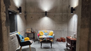 A cozy room with a concrete wall, a window, a black chair, a colorful sofa, a small table, and a cabinet. - The Mill Hotel (Fergus Falls)