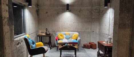 Une pièce chaleureuse avec un mur en béton, une fenêtre, une chaise noire, un canapé coloré, une petite table et un meuble.