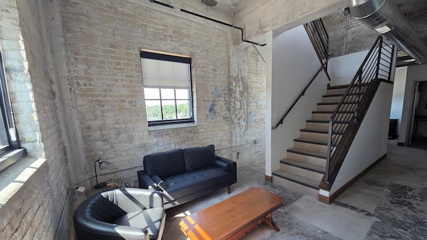 A spacious loft with exposed brick walls, a black leather sofa, a wooden coffee table, and a staircase. - The Mill Hotel (Fergus Falls)