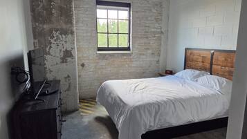 Une chambre avec un lit simple, une tête de lit en bois, une table de chevet avec une horloge, un téléviseur posé sur une commode et une fenêtre donnant sur un paysage verdoyant.