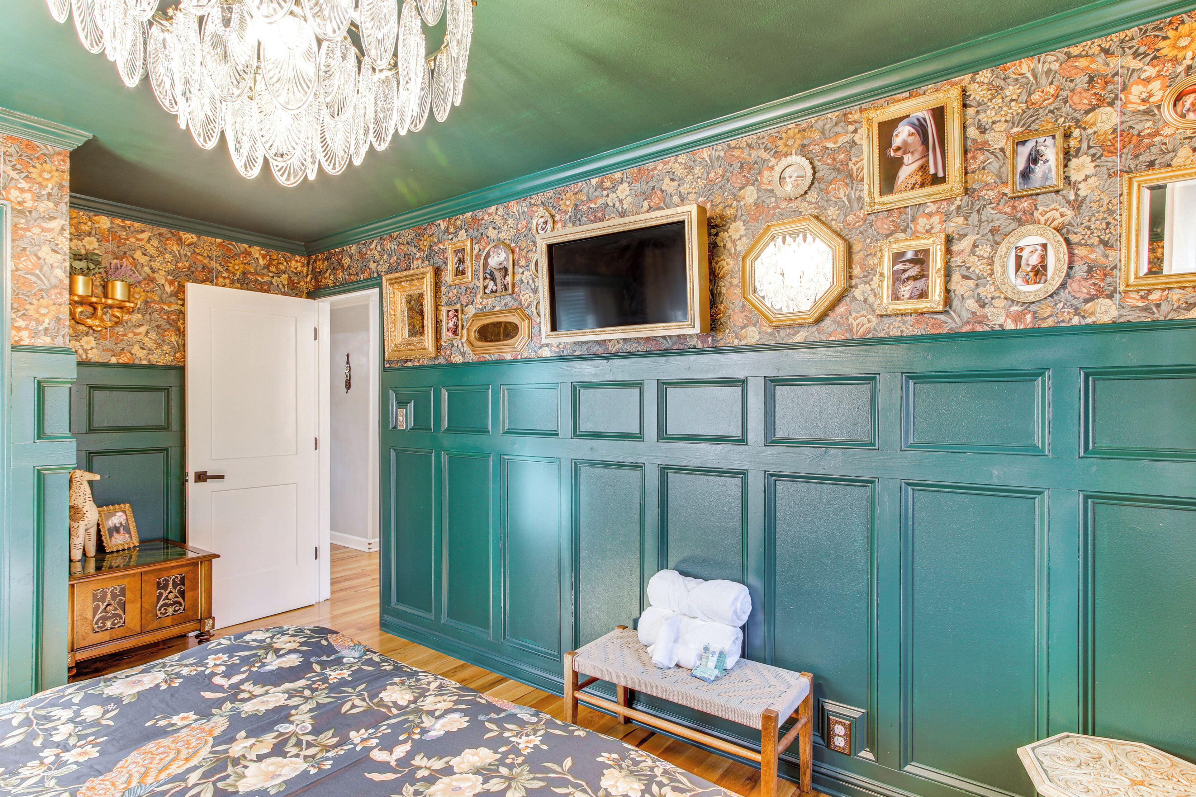A room with a bed, a bench, a television, and a wall decorated with framed pictures and a floral pattern.