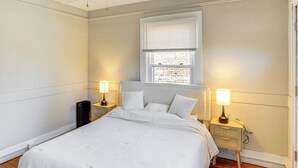 A bedroom with a large bed, two bedside tables with lamps, a window with blinds, and a radiator. - Walk to Dtwn Hudson: Cozy Upstate Apartment (Hudson)