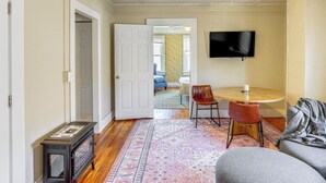 A room with a wooden floor, a patterned rug, a table with chairs, a television mounted on the wall, and a door leading to another room. - Walk to Dtwn Hudson: Cozy Upstate Apartment (Hudson)