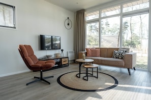 A modern living room with a sofa, armchair, TV, and a large window. - Luxury chalet with hot tub in wooded area of the Sallandse Heuvelrug (Haarle)