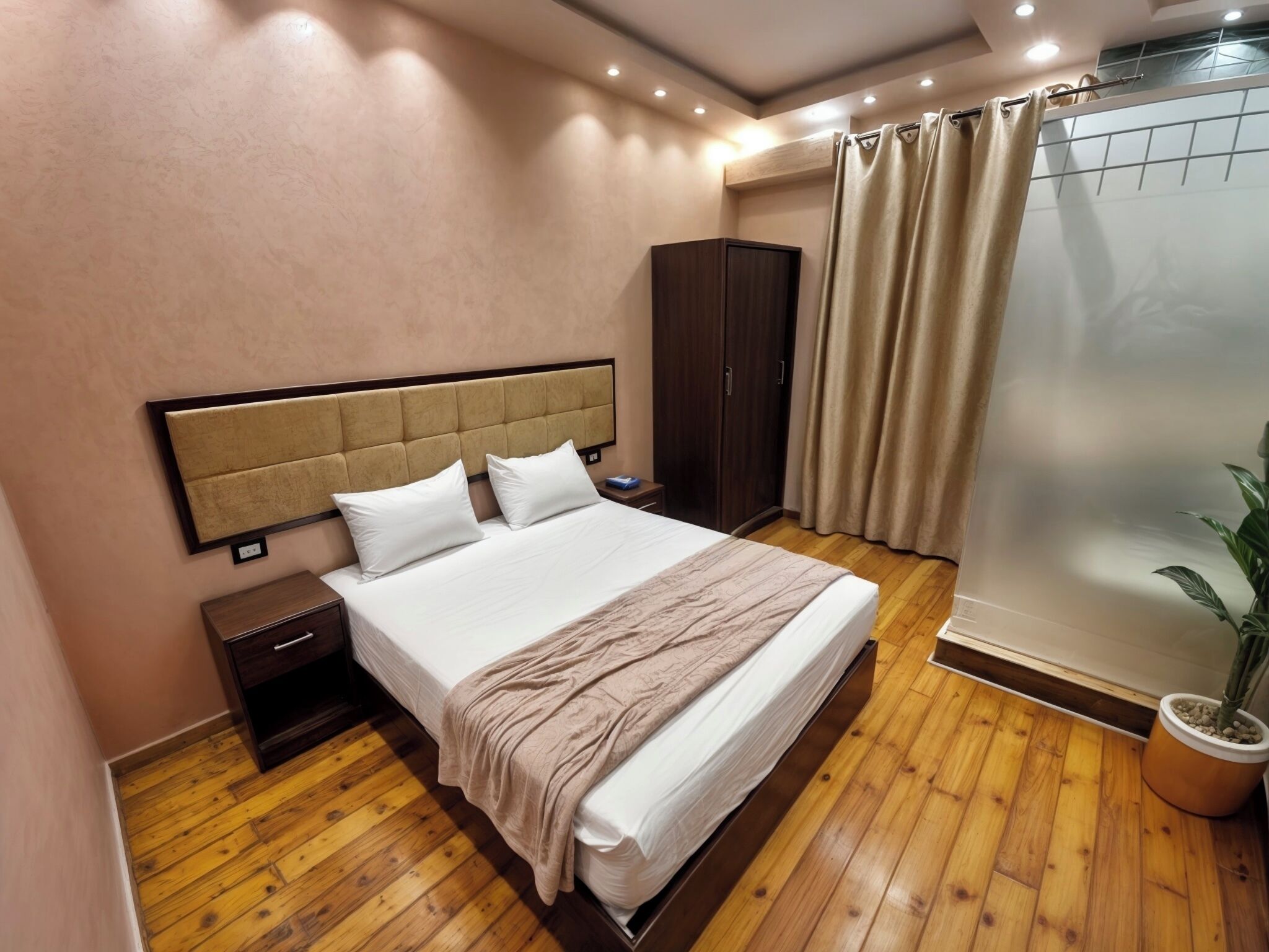 A hotel room with a large bed, wooden flooring, a wardrobe, a nightstand, and a potted plant.