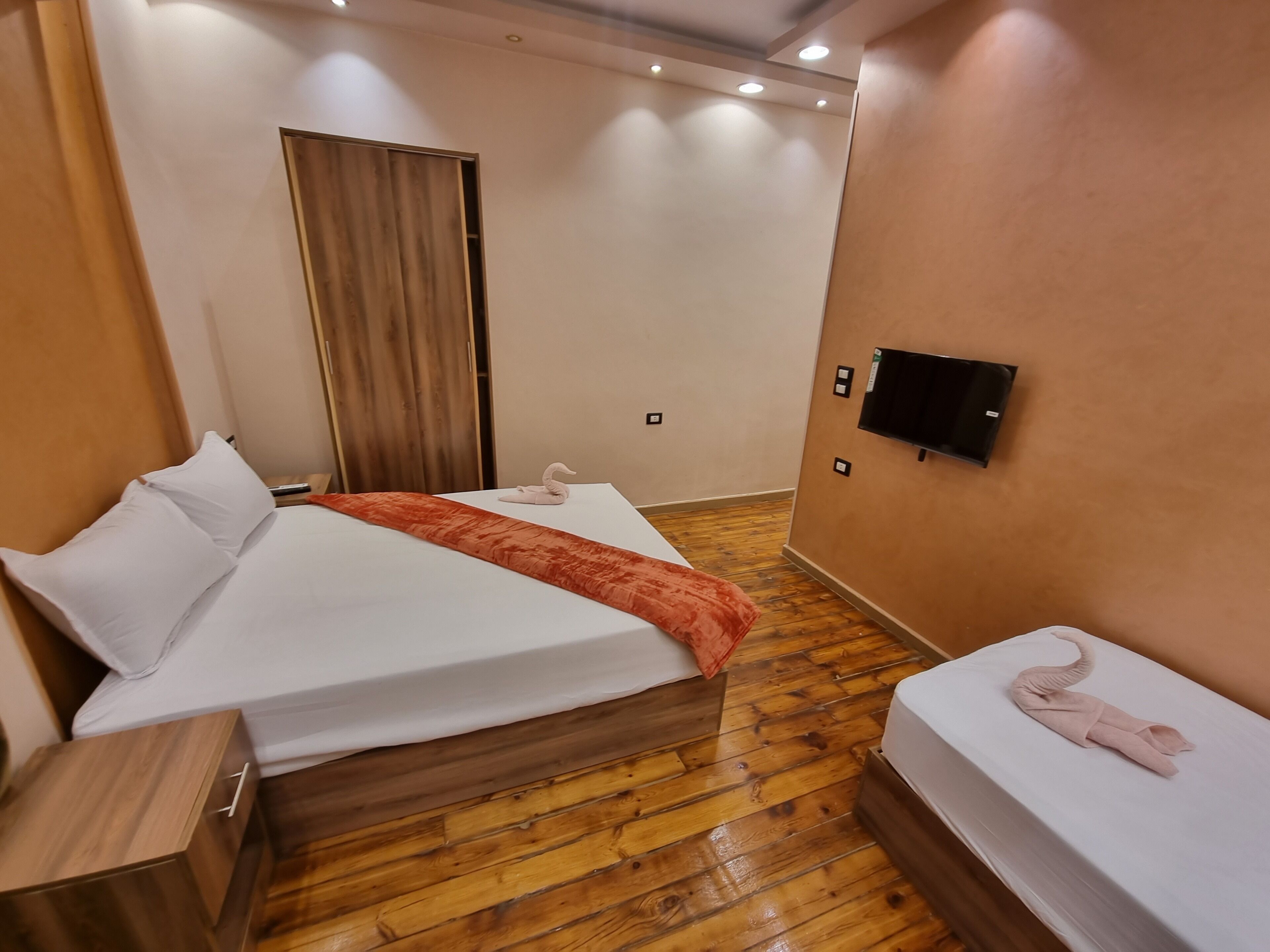 Habitación de hotel con piso de madera, cama con sábanas blancas, armario de madera, televisor de pantalla plana en la pared y cortina en la puerta.