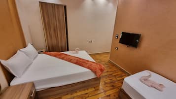 A hotel room with a wooden floor, a bed with white linens, a wooden wardrobe, a flat-screen TV mounted on the wall, and a curtain on the door.