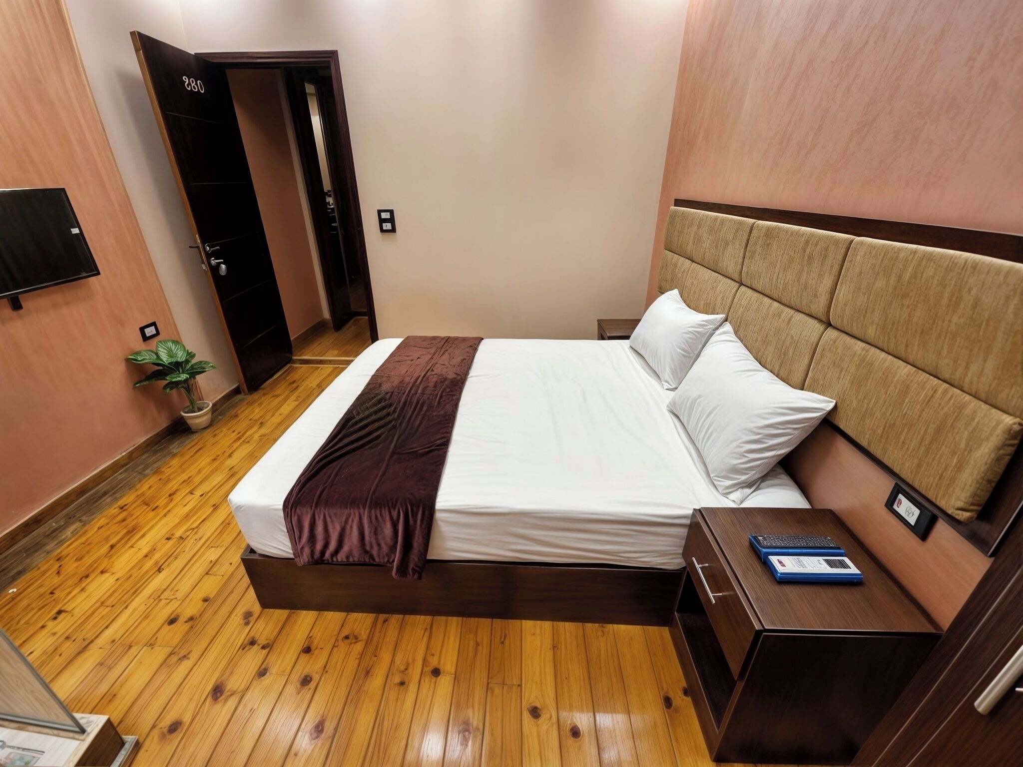 Una habitación de hotel con piso de madera, una cama con sábanas blancas y un plaid marrón, una mesita de noche con una computadora portátil y una planta en maceta.