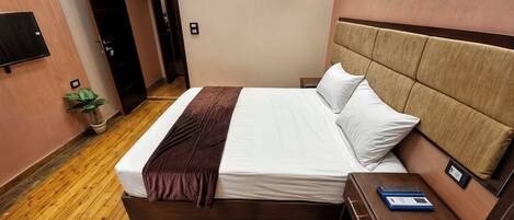 Una habitación de hotel con piso de madera, una cama con sábanas blancas y un plaid marrón, una mesita de noche con una computadora portátil y una planta en maceta.