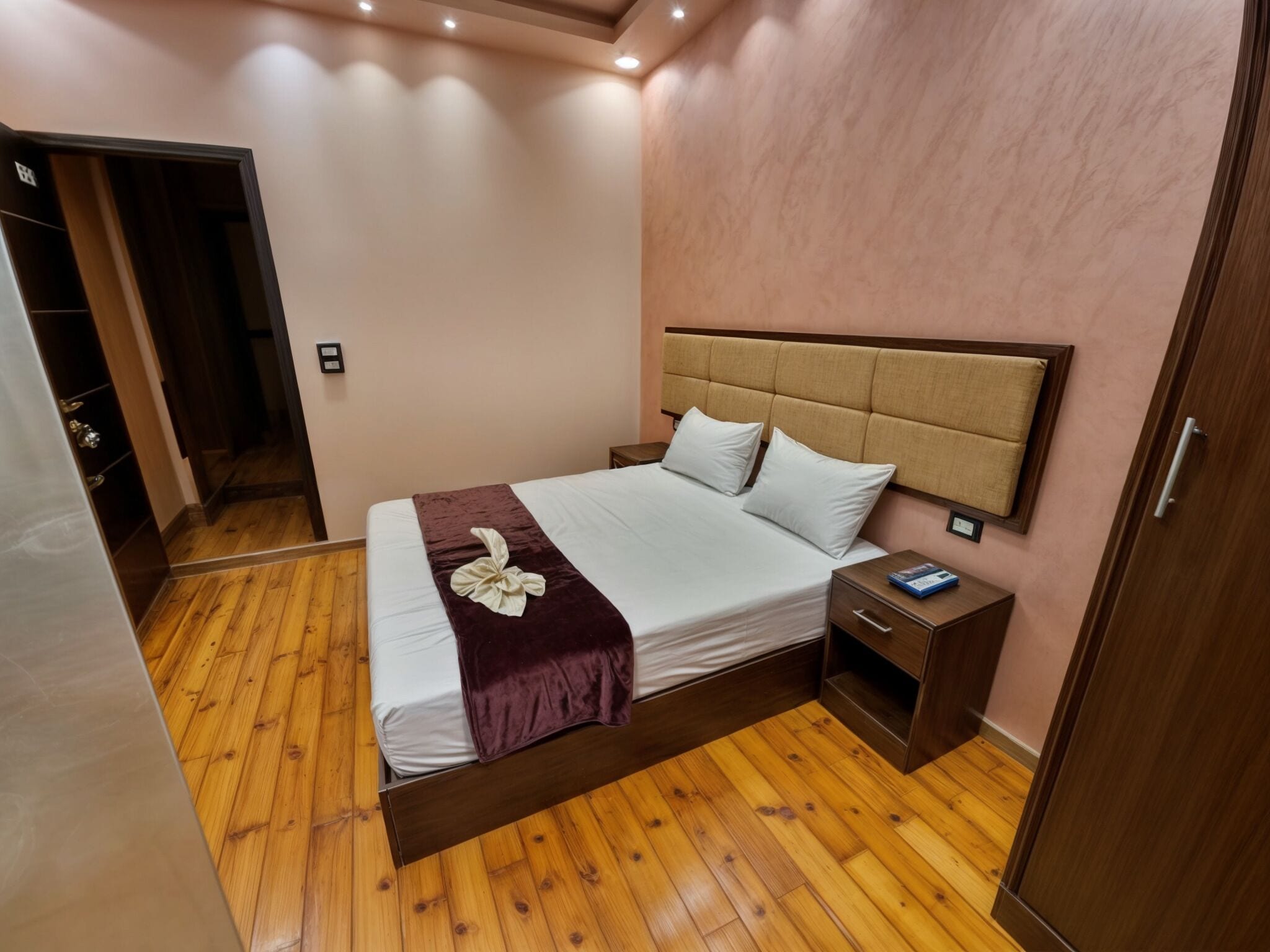 Un dormitorio ordenado con piso de madera, una cama con sábanas blancas, un cabecero marrón y una mesita de noche con un libro.