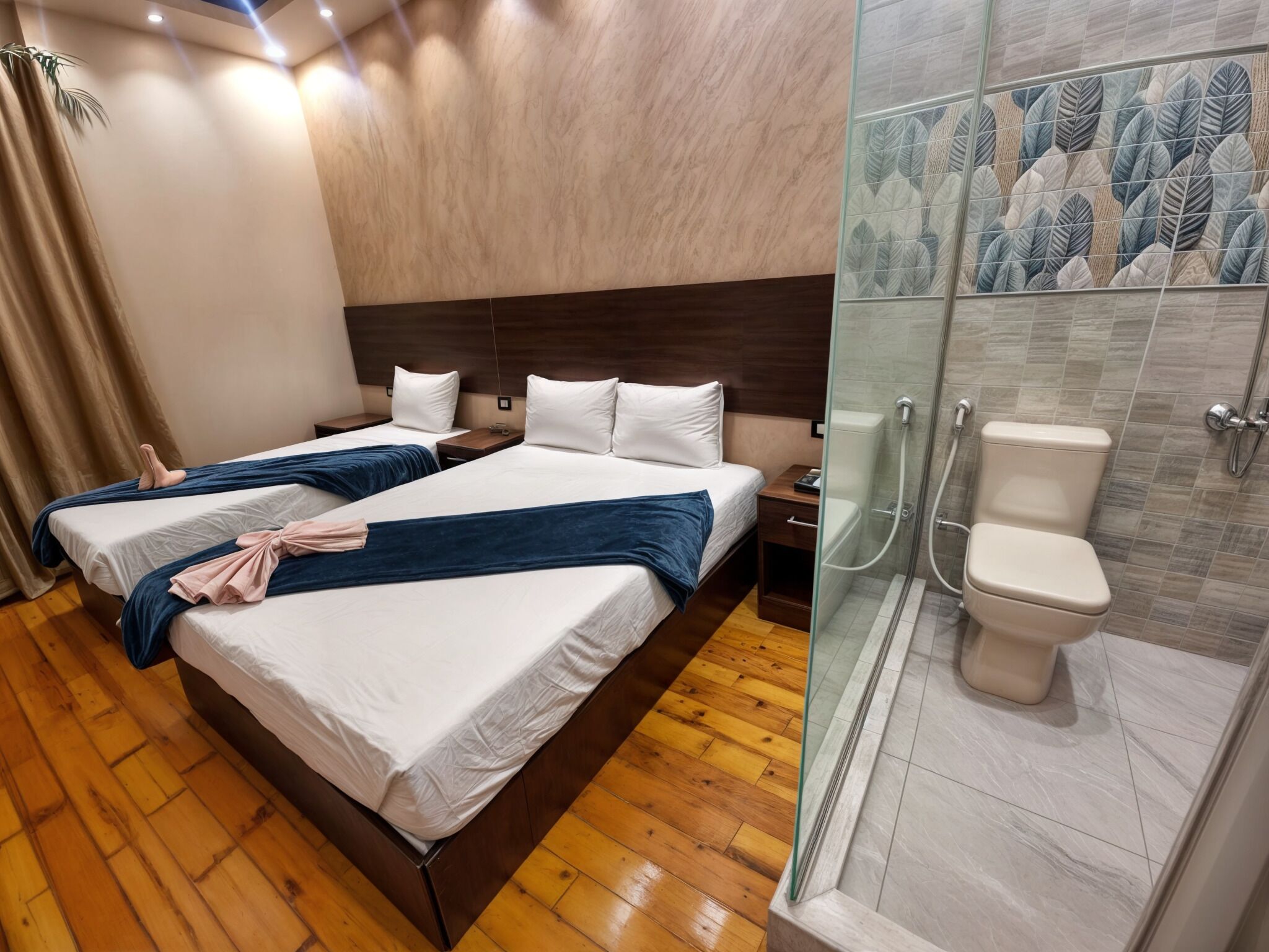 A hotel room with two beds, wooden flooring, a shower with a glass door, and a toilet.