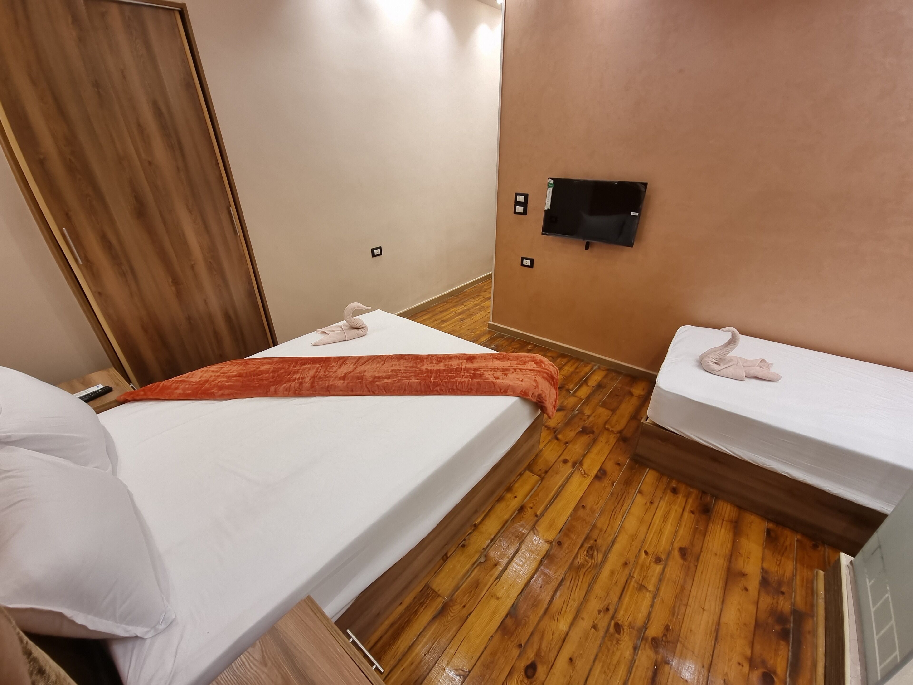 A hotel room with two beds, wooden flooring, a TV mounted on the wall, and a wooden door.