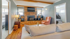 Un salon chaleureux avec une cheminée en briques, un téléviseur fixé au-dessus, et un coin salon confortable avec un canapé et des fauteuils.