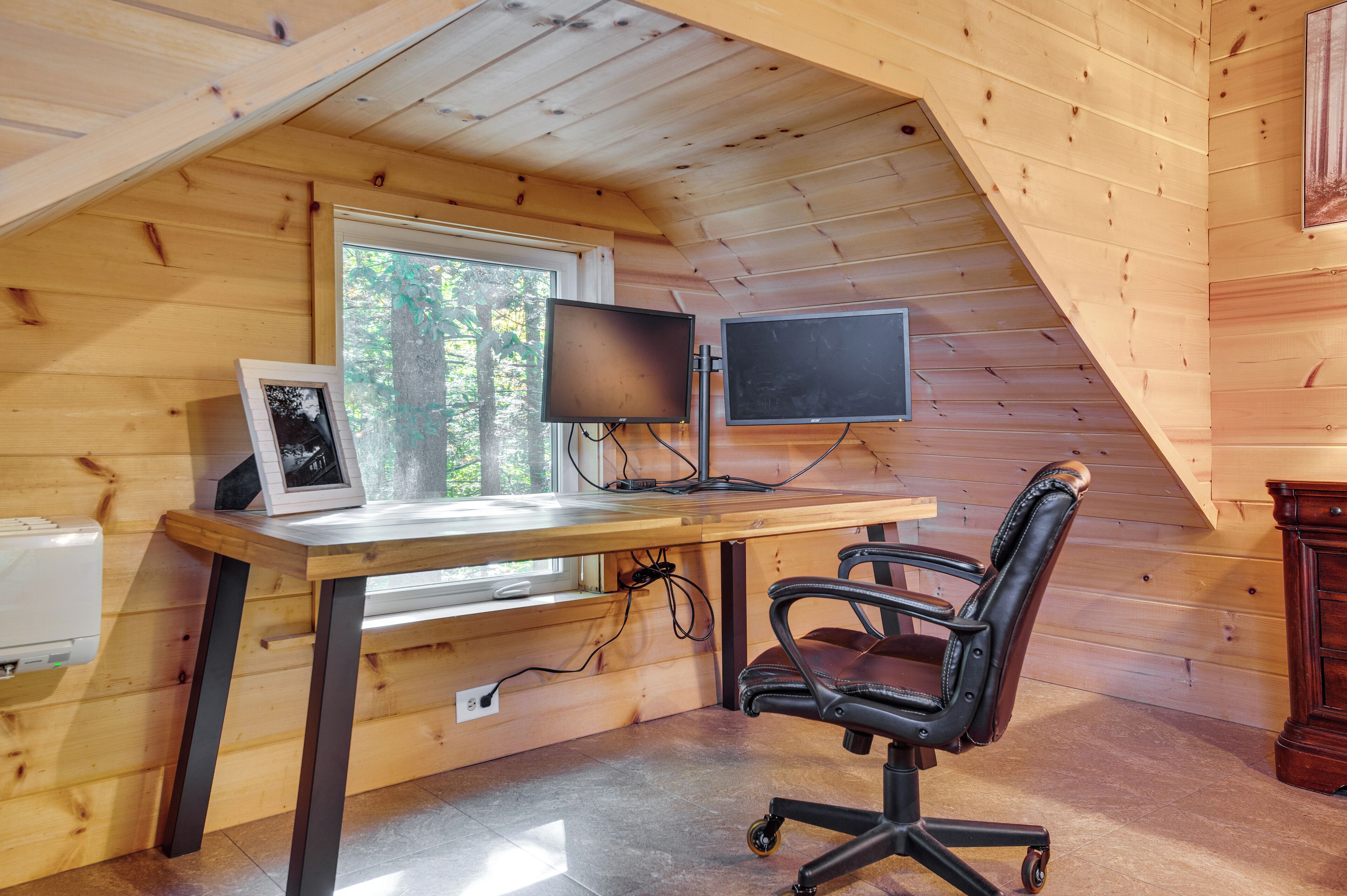A wooden-paneled room with a desk, two monitors, a chair, and a window overlooking trees.