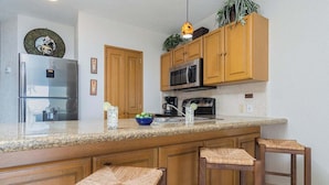Eine Küche mit Holzschränken, einem Edelstahlkühlschrank, einer Mikrowelle und einer Arbeitsplatte aus Granit. Drei Hocker mit Sitzgeflecht sind vorhanden.